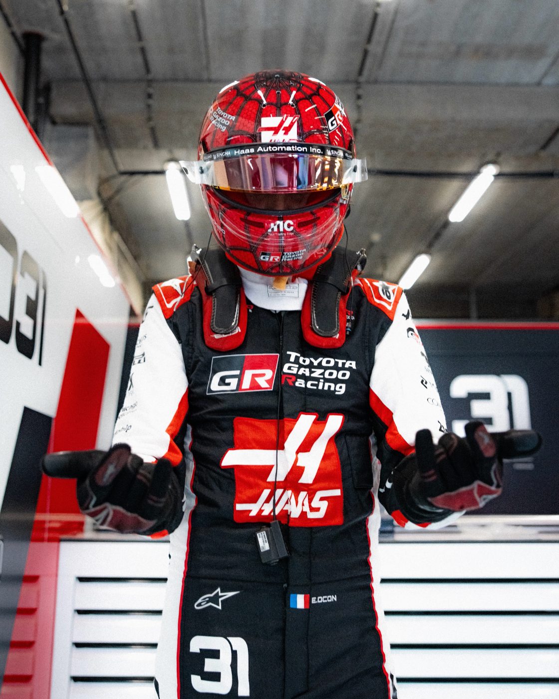 Esteban Ocon debout dans le garage Haas, casque Spider-Man, bras ouverts
